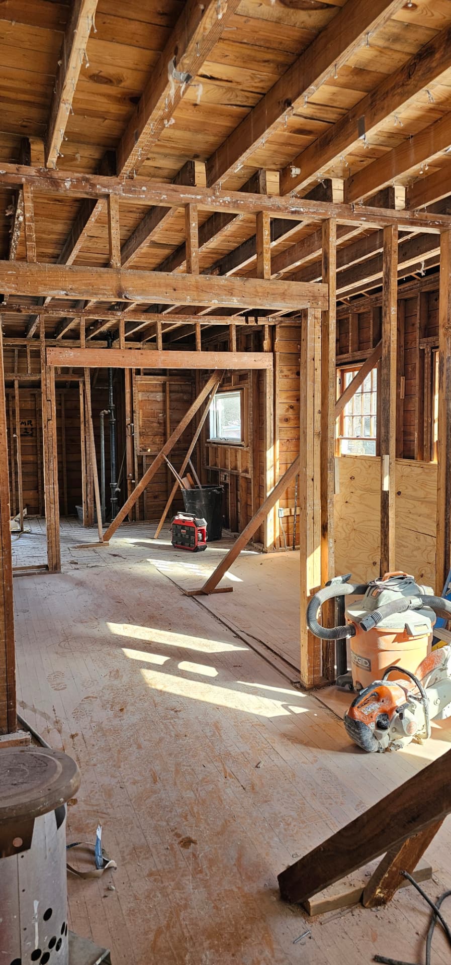 Opened-up upper floor with exposed rafters, stud walls, and sunlit subfloor during renovation