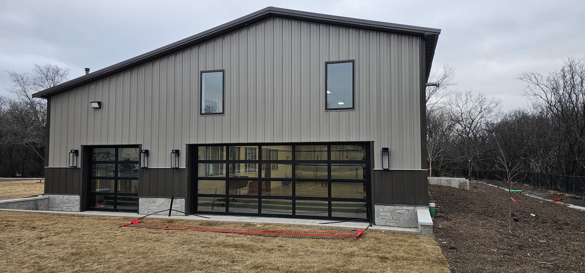 Gray metal building with two black-framed glass overhead doors, upper windows, and wall lights