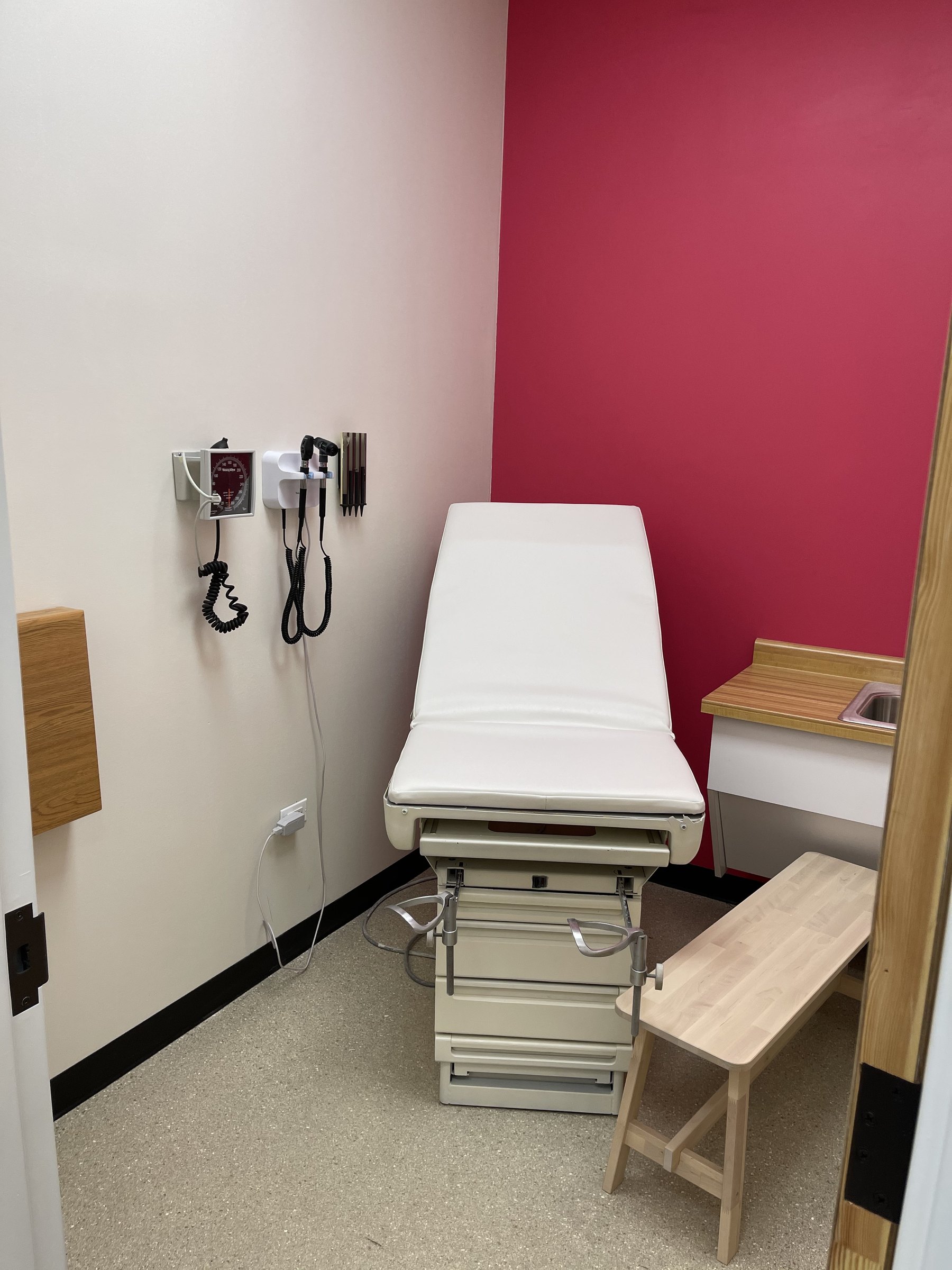 Finished exam room with exam table, red accent wall, wall-mounted diagnostic equipment, and sink counter
