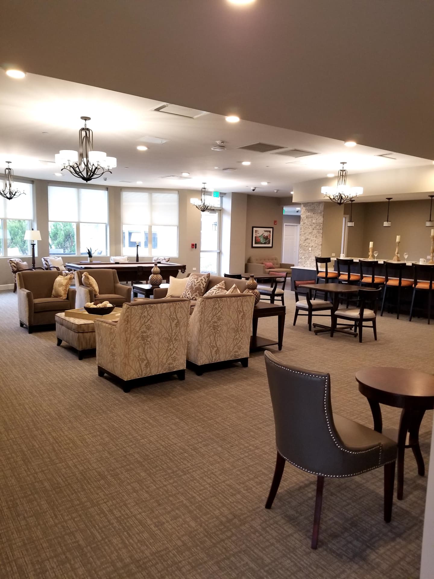 Open common lounge with armchairs around an ottoman, a pool table, and a bar with barstools